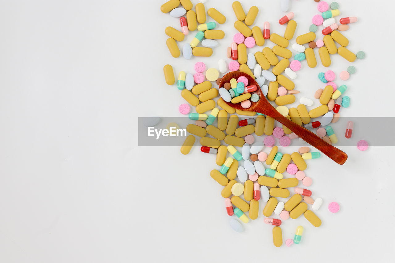 High angle view of multi colored pills with wooden spoon on white background