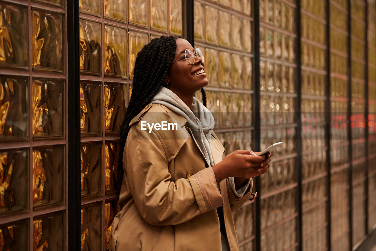 Confident black female with braids in casual outfit looking away and browsing smartphone while standing near glass wall