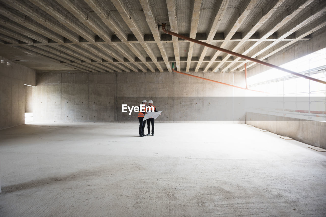 Two men with plan wearing safety vests talking in building under construction