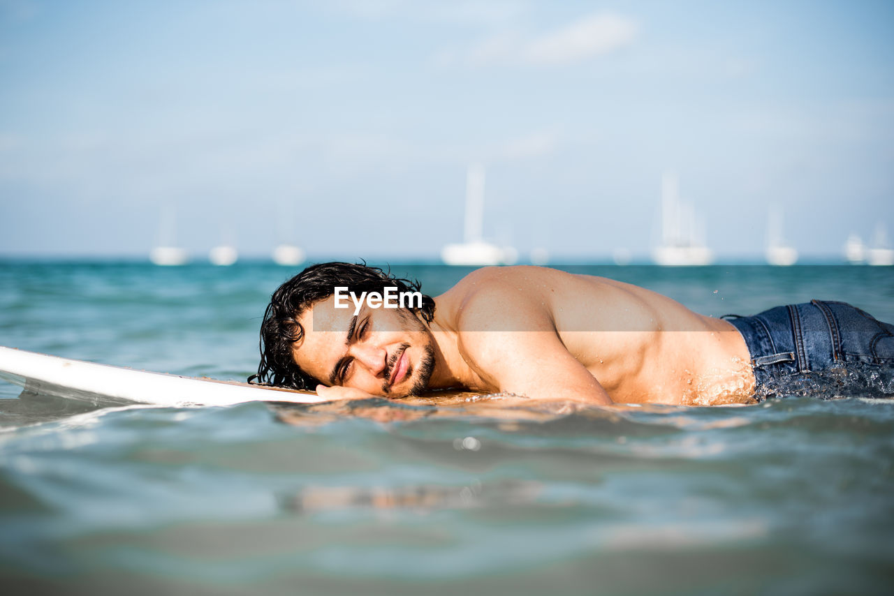 PORTRAIT OF SHIRTLESS MAN RELAXING AT SEA AGAINST SKY