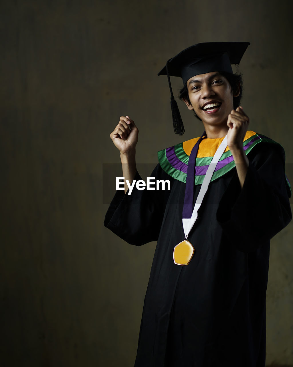 Portrait of smiling man wearing graduation gown standing against wall