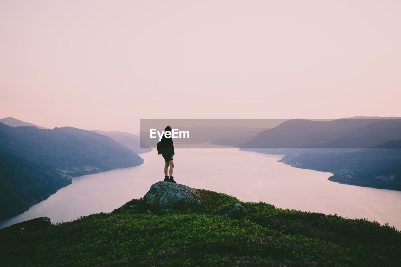 Rear view of woman looking at river amidst mountains against sky during sunset