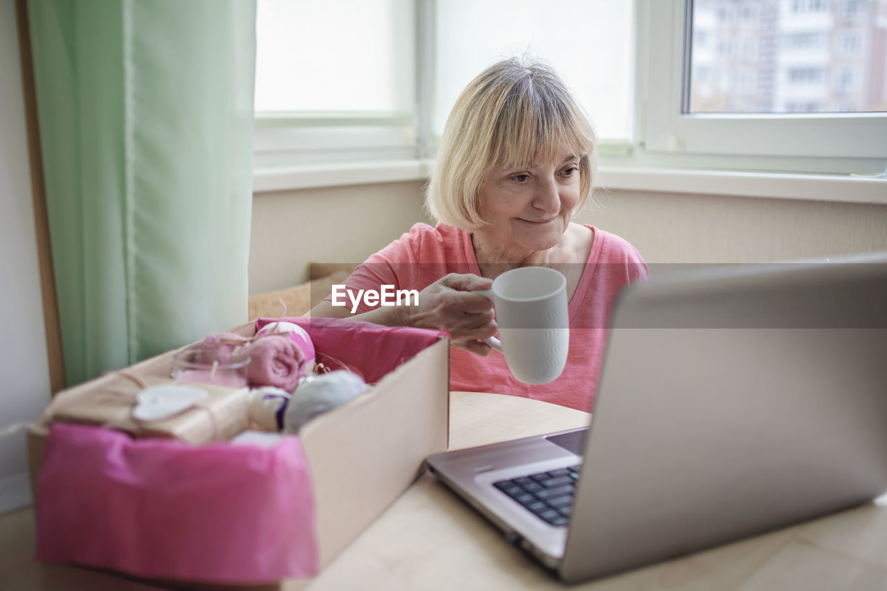Senior woman holding coffee cup using laptop at home