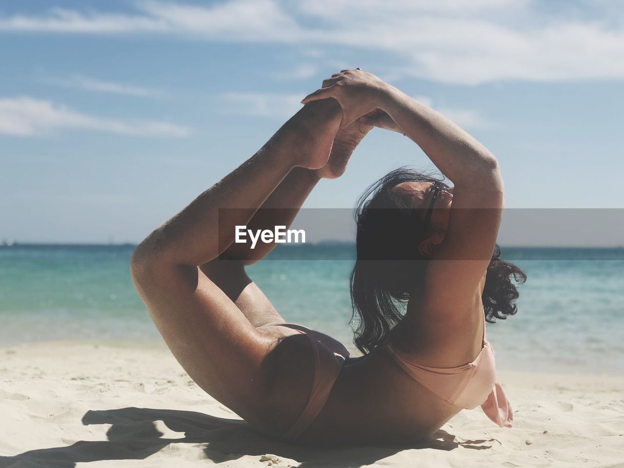 Side view of woman wearing bikini practicing yoga while lying on sand at beach against sky during sunny day