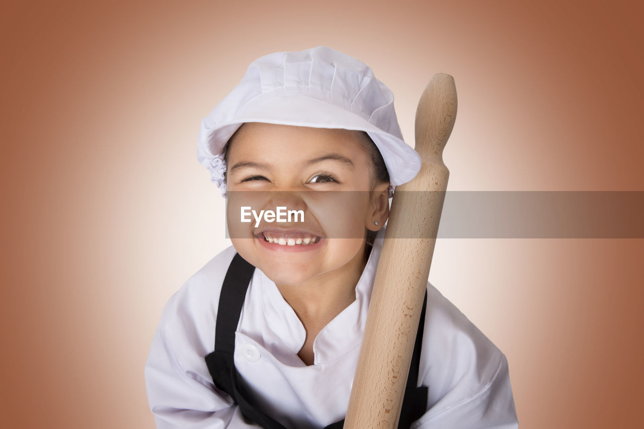 Portrait of girl in chef uniform holding rolling pin against orange background