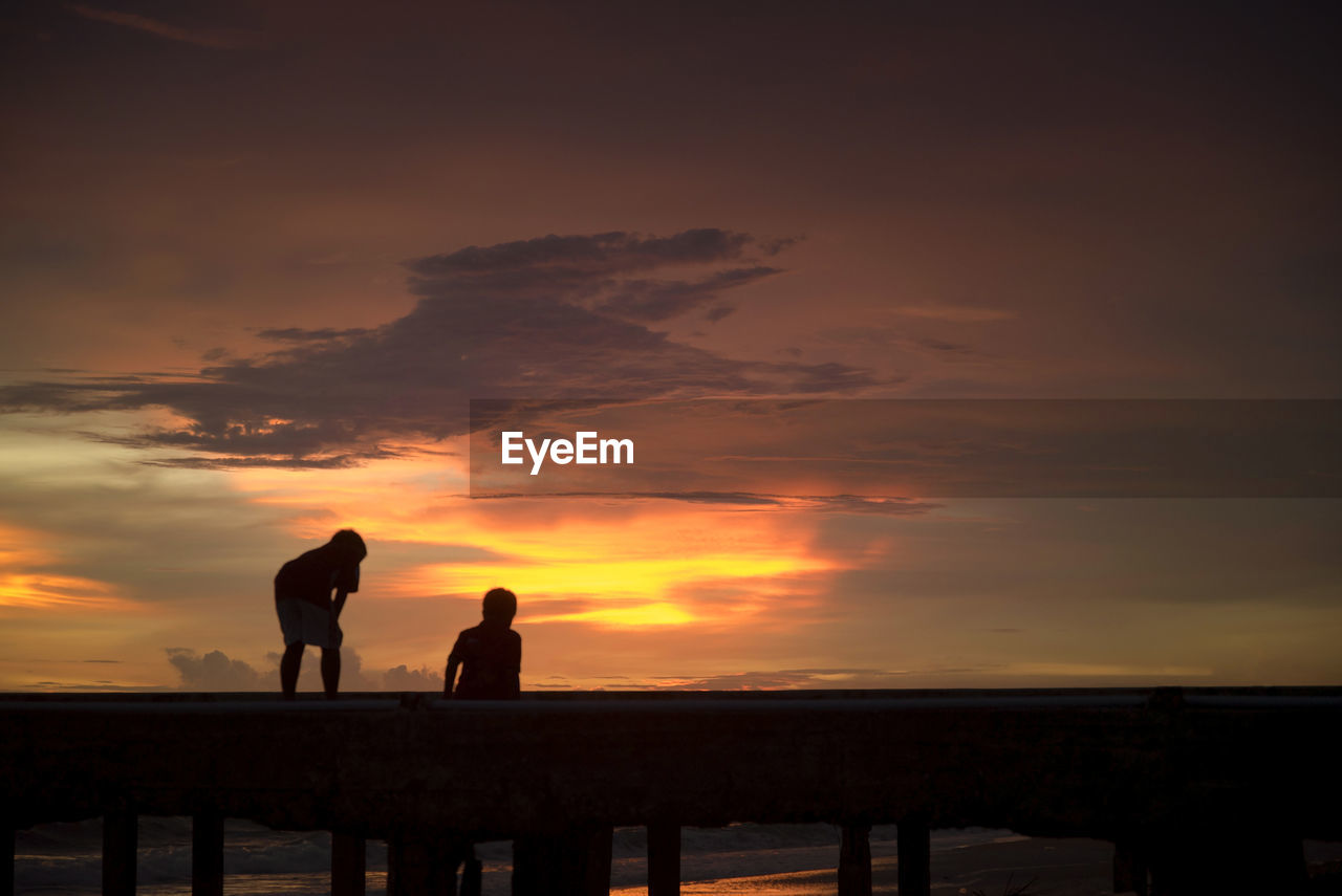 SILHOUETTE MEN STANDING BY SEA AGAINST SKY DURING SUNSET