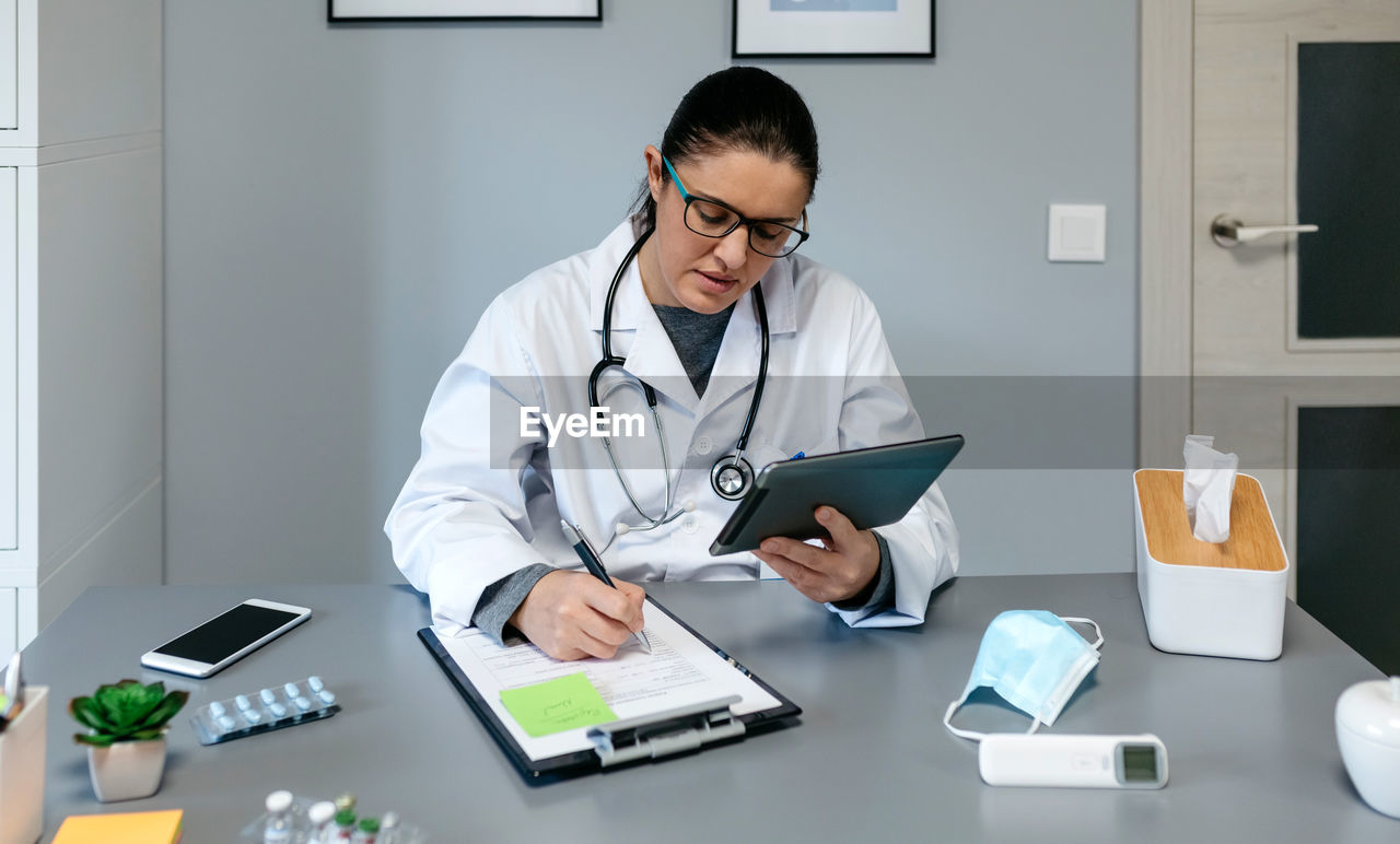 Doctor using digital tablet while writing on document at desk in office