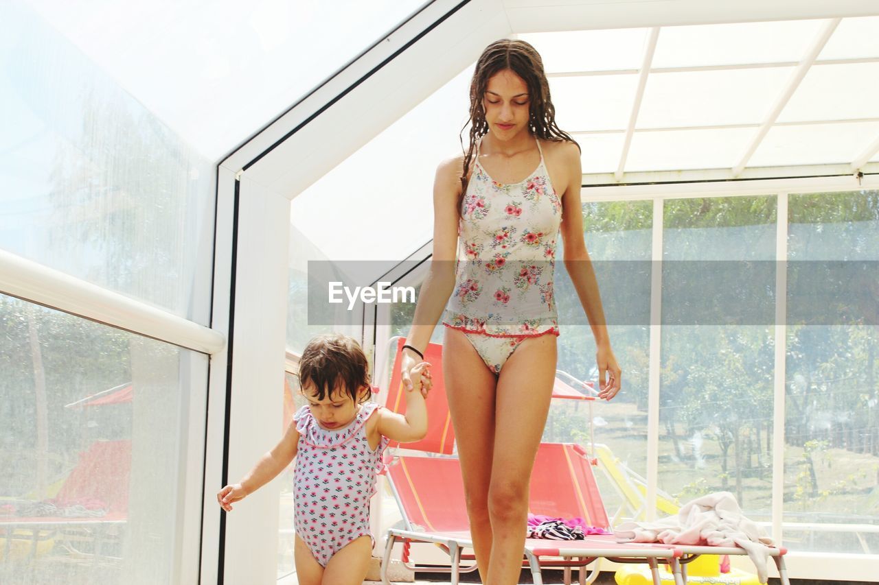 Mother and daughter wearing swimwear while standing by window at home