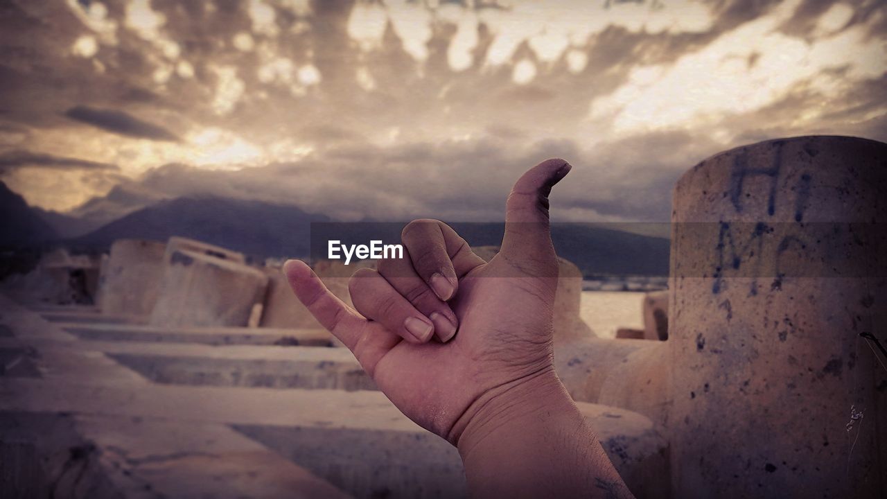 Cropped image of hand showing shaka sign against cloudy sky during sunset