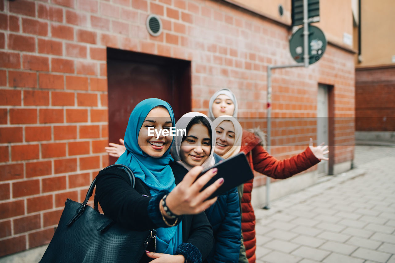 Happy woman taking selfie with female friends on sidewalk against building in city