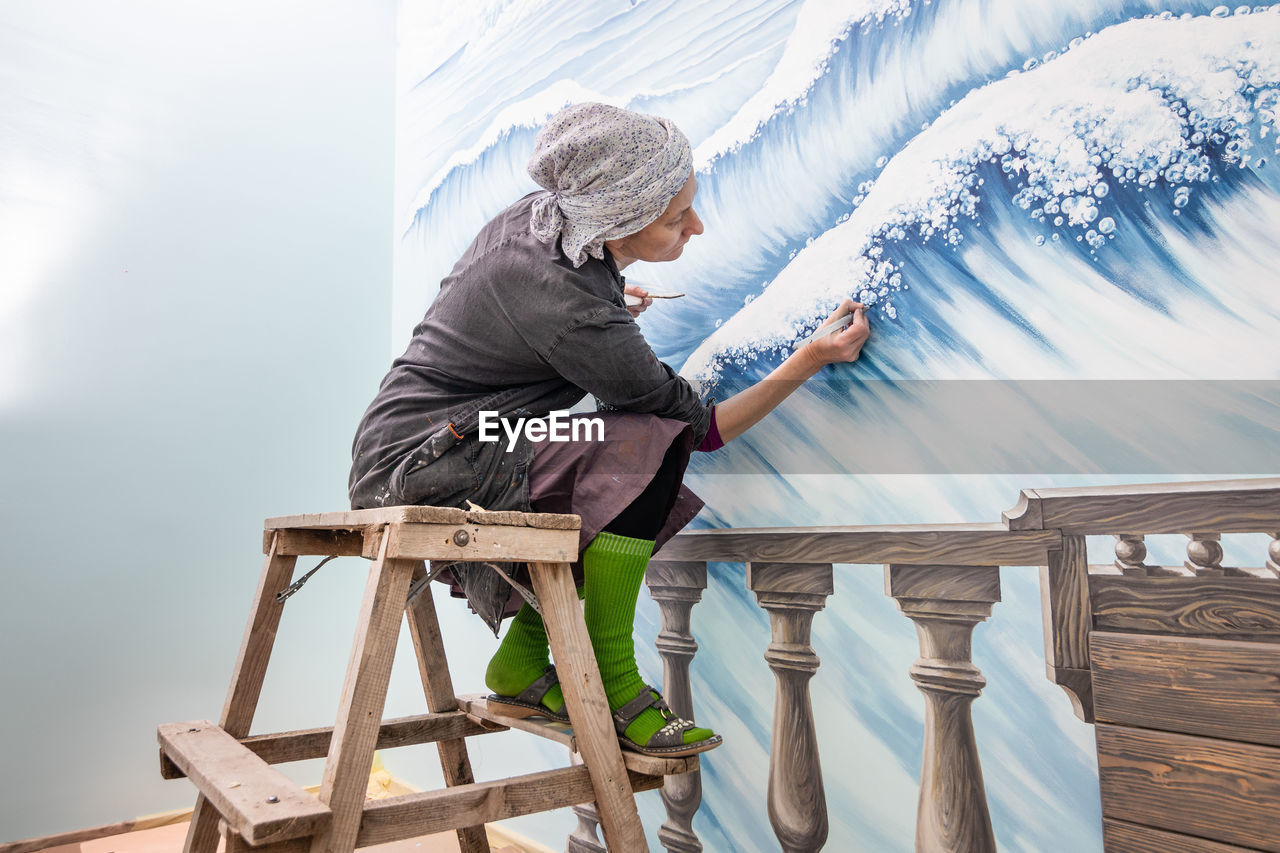 Mature woman artist draws a mural on a marine theme sitting on a ladder