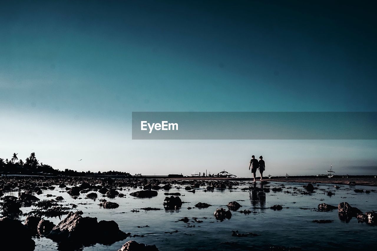 Silhouette couple walking at beach against blue sky during sunset