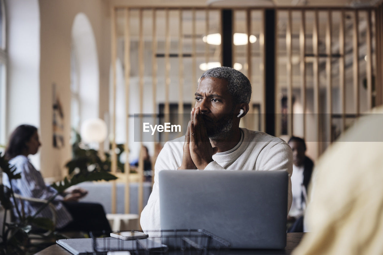 Worried businessman with hands clasped talking through wireless in-ear headphones at office