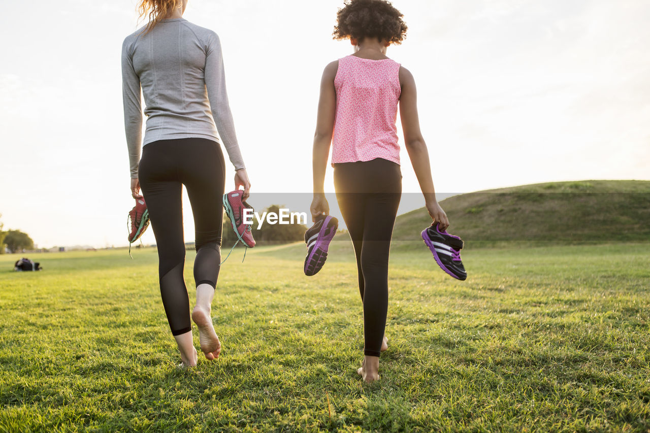 Rear view of mother and daughter holding sports shoes while walking on grass at park during sunset