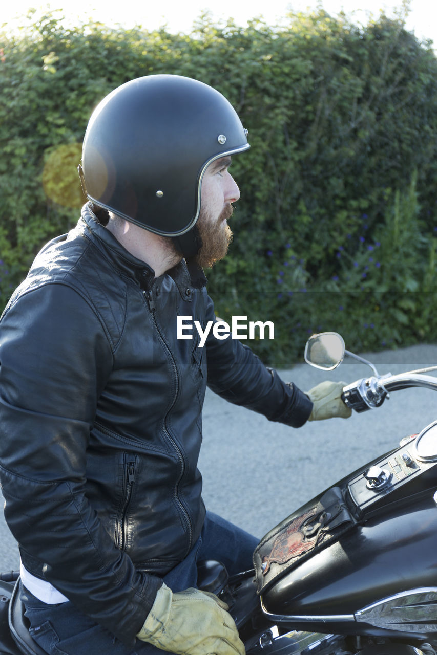 Side view of biker looking away while sitting on motorcycle against plants during sunset