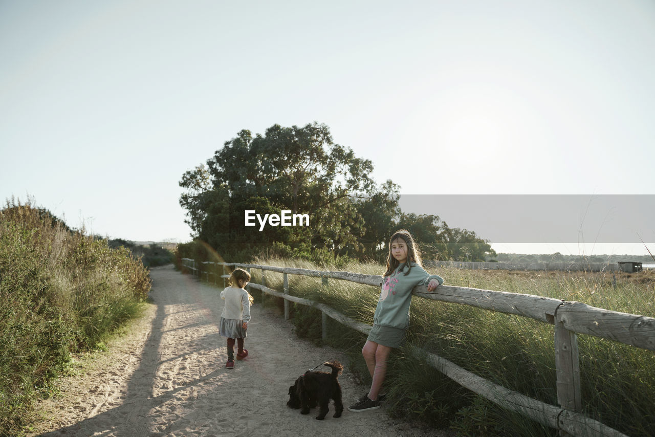 Rear view of two girl walking on field with dog against clear sky