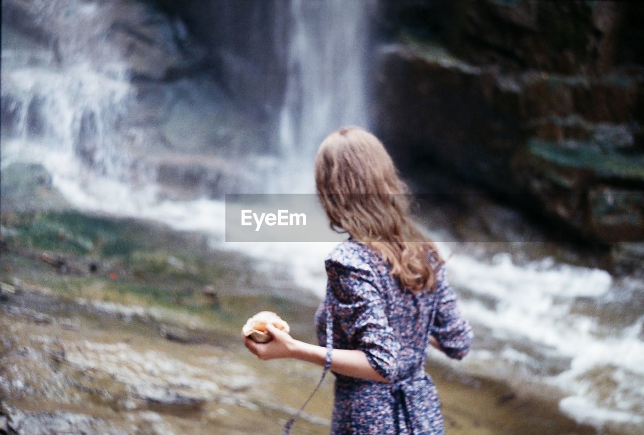 Rear view of girl holding food while looking at waterfall