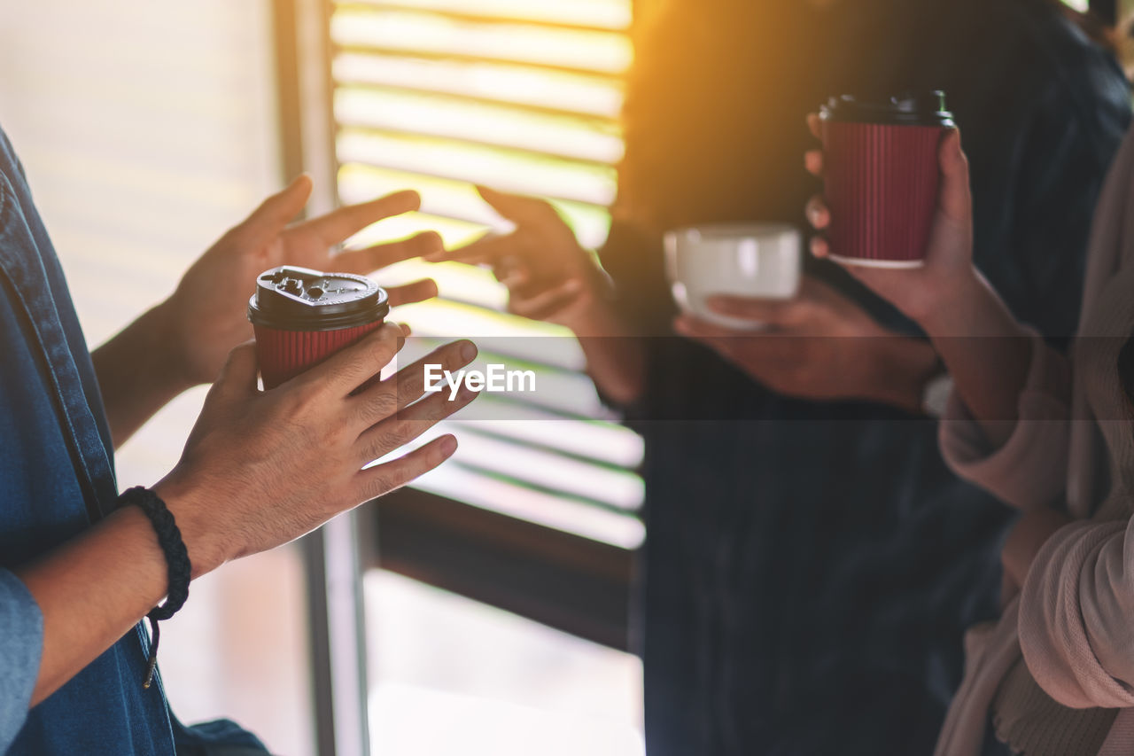 Midsection of people holding disposable coffee cup while discussing in office