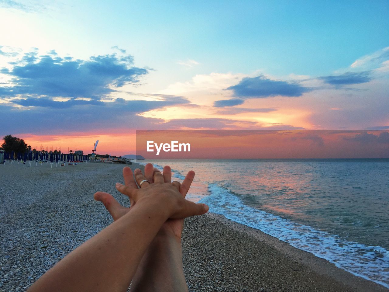 Cropped hands of person by sea against sky during sunset