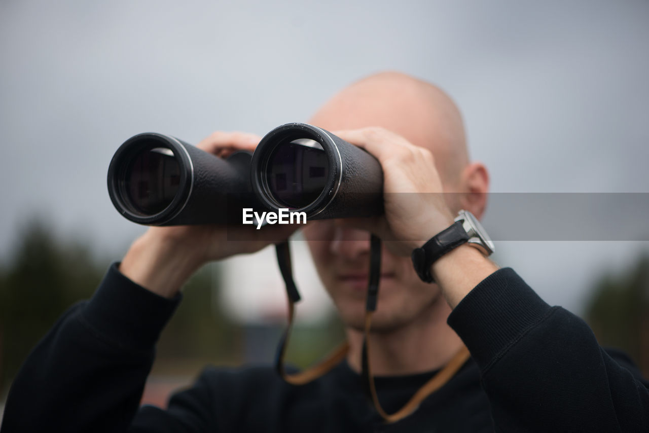 Close-up of mid adult man looking through binoculars while standing against sky