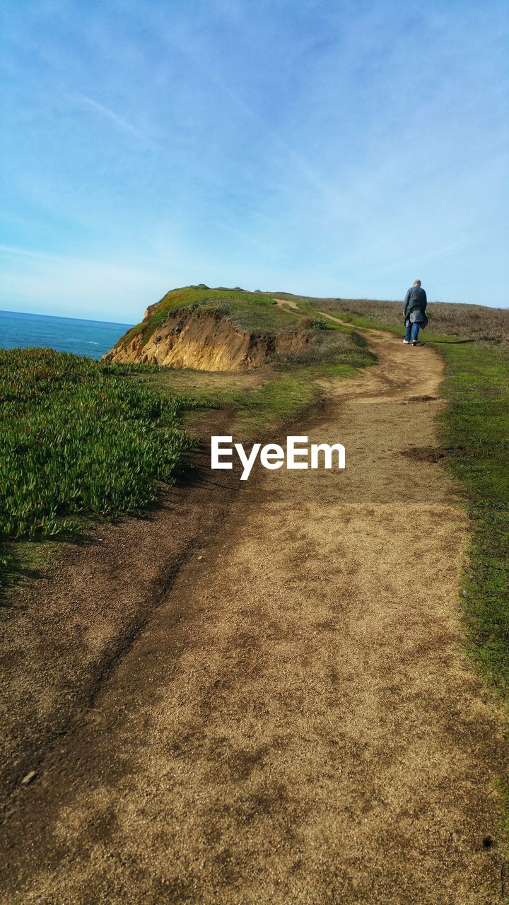 Rear view of man on sandstone hiking path at ocean cliff's edge against sky