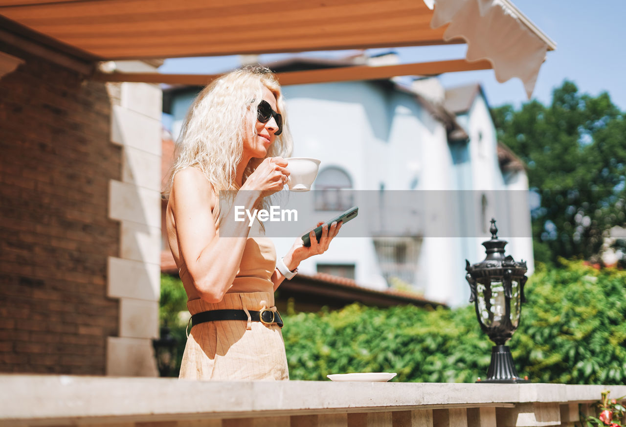 Adult elegant blonde woman in sunglasses and beige clothes using mobile phone at house backyard