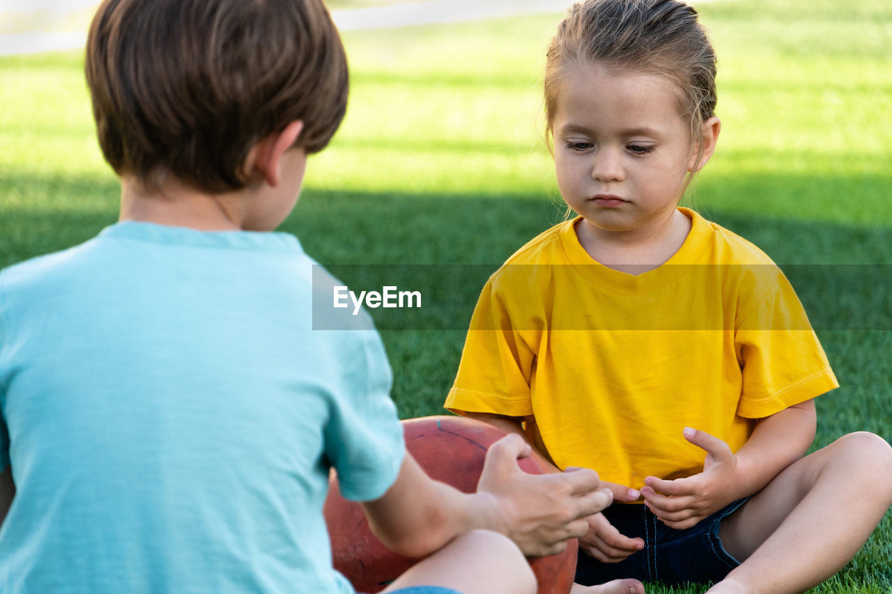 Cute little girl sitting on lawn with brother, talking with him and looking to ball