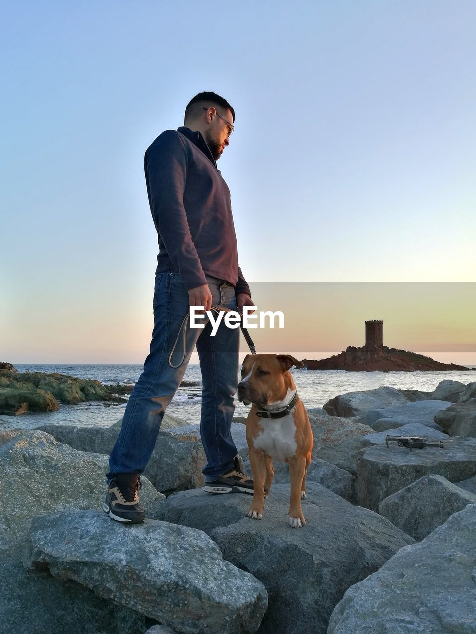 Man with dog standing on rock against sky during sunset