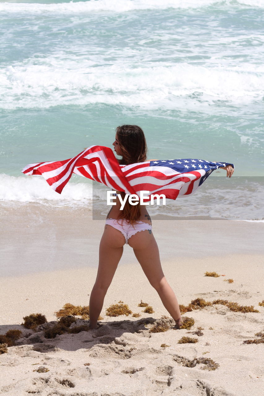 Rear view of woman standing with american flag at beach