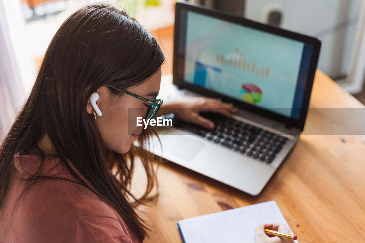 Happy young female in wireless earbuds and eyeglasses sitting at table with cup of hot drink and typing on modern netbook at home