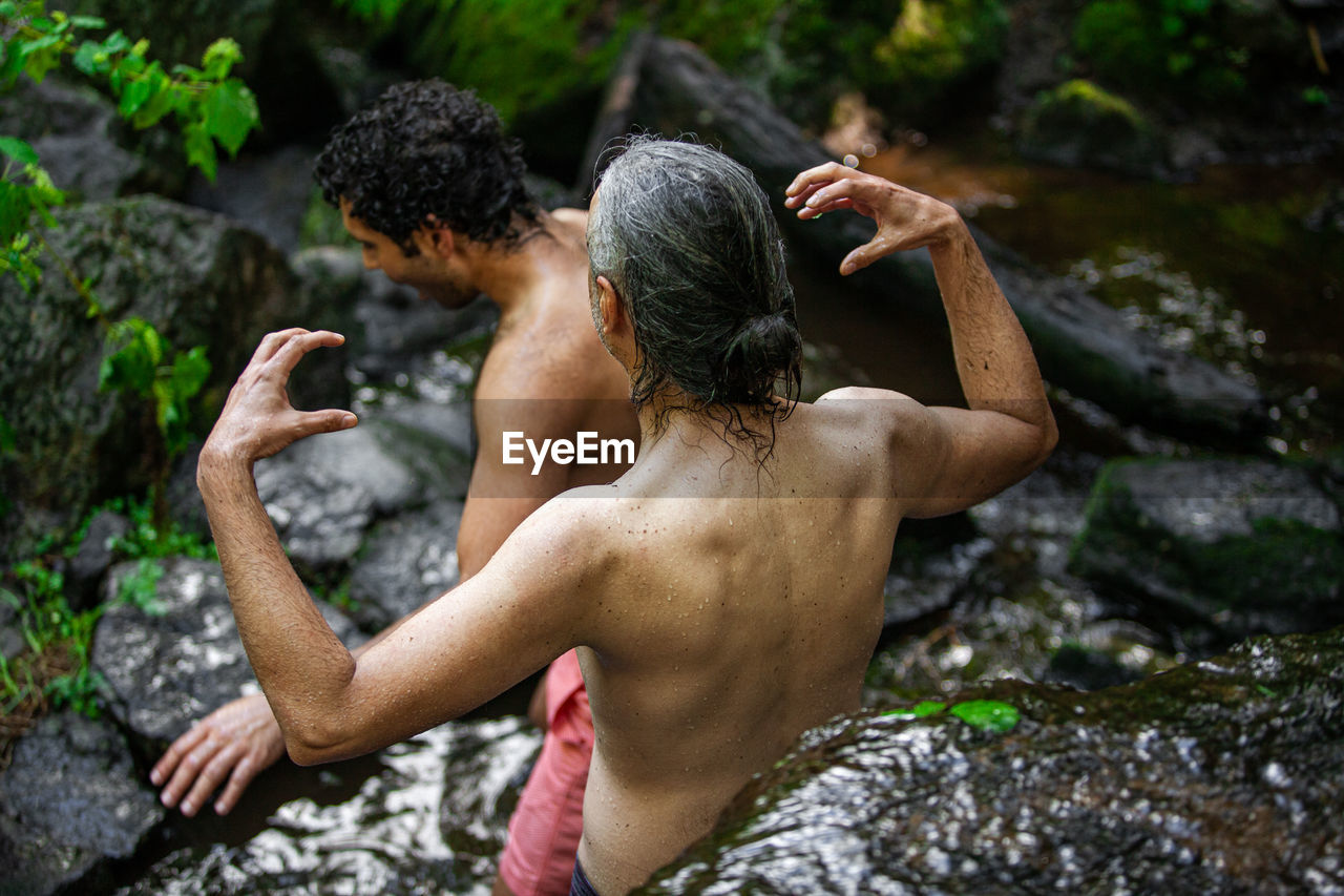 REAR VIEW OF SHIRTLESS MAN SITTING ON ROCKS