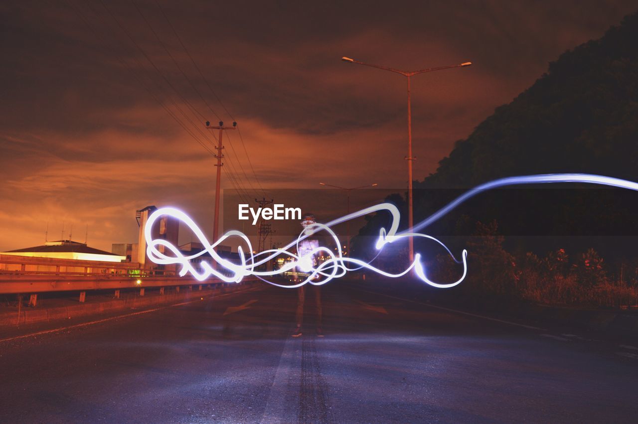 Young man with illuminated light painting standing on road against sky at night