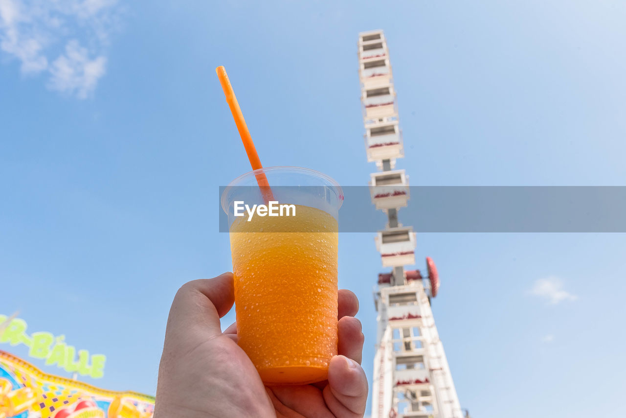 Cropped hand holding juice glass against ferris wheel in city