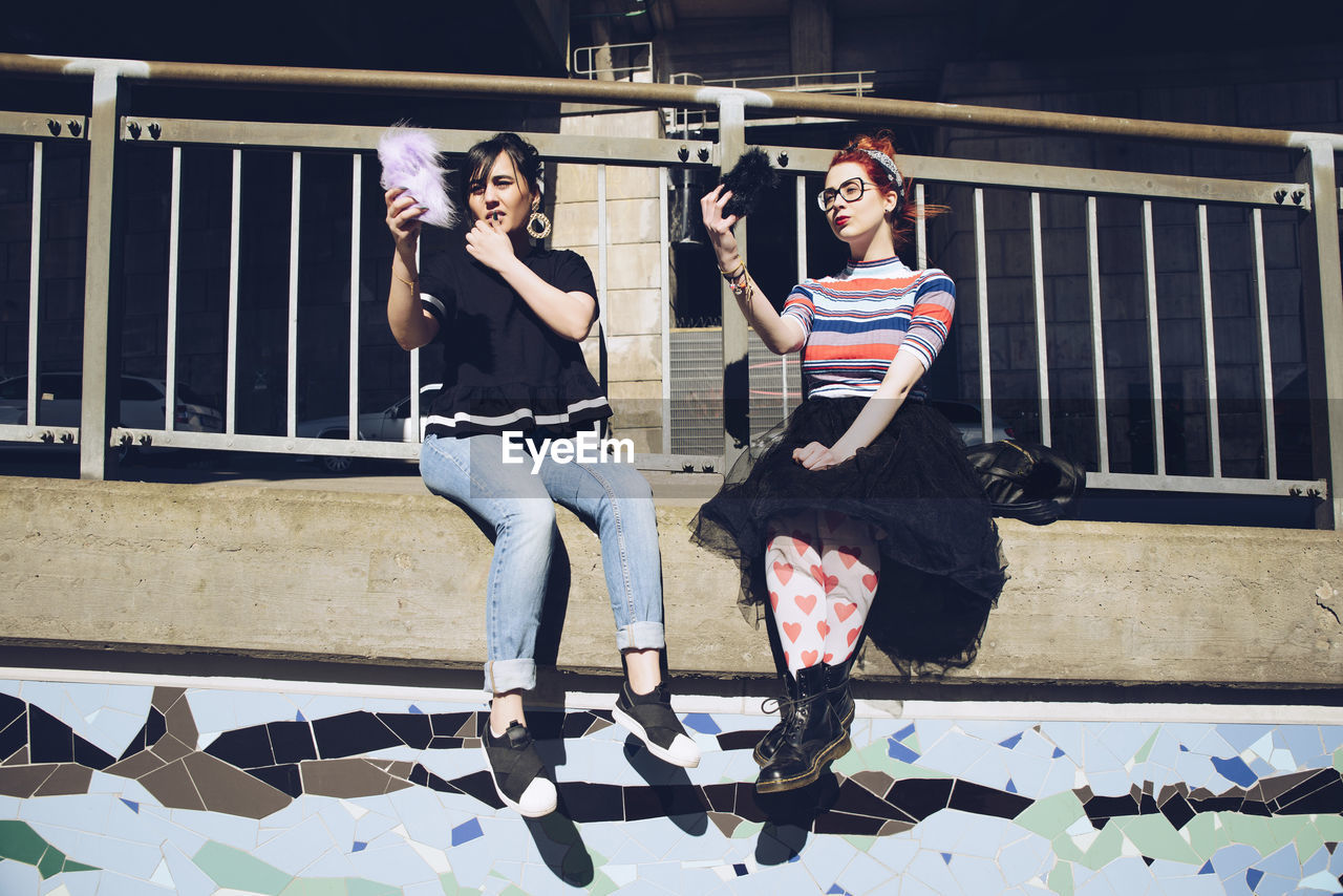 Hipster female friends taking selfie with mobile phones while sitting on wall against railing