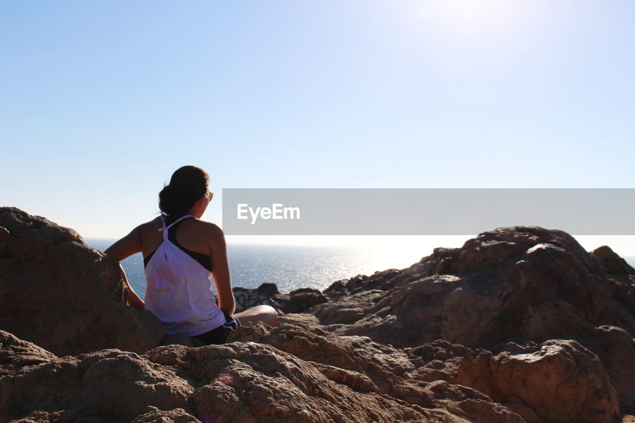 Rear view of woman sitting on rocks by sea against clear sky