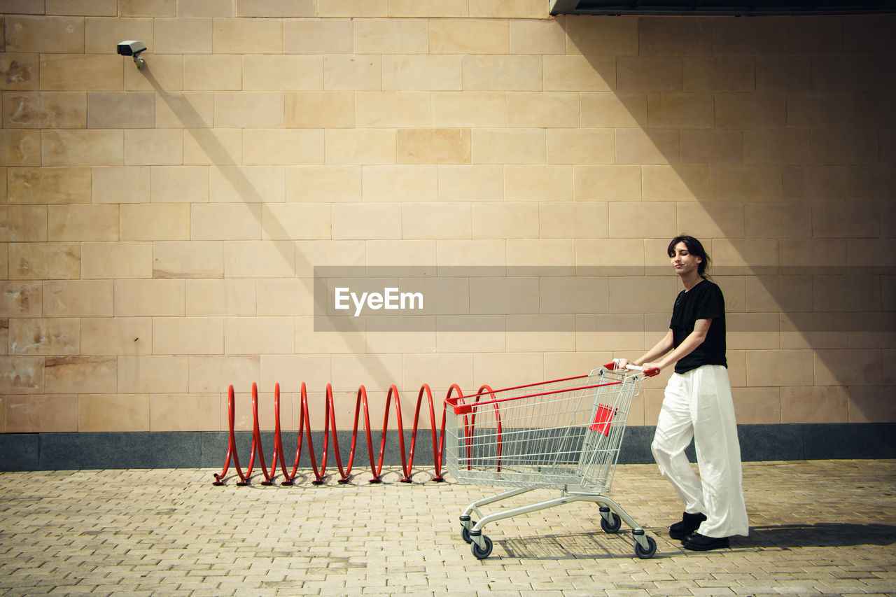 Full length of woman pushing shopping cart against wall