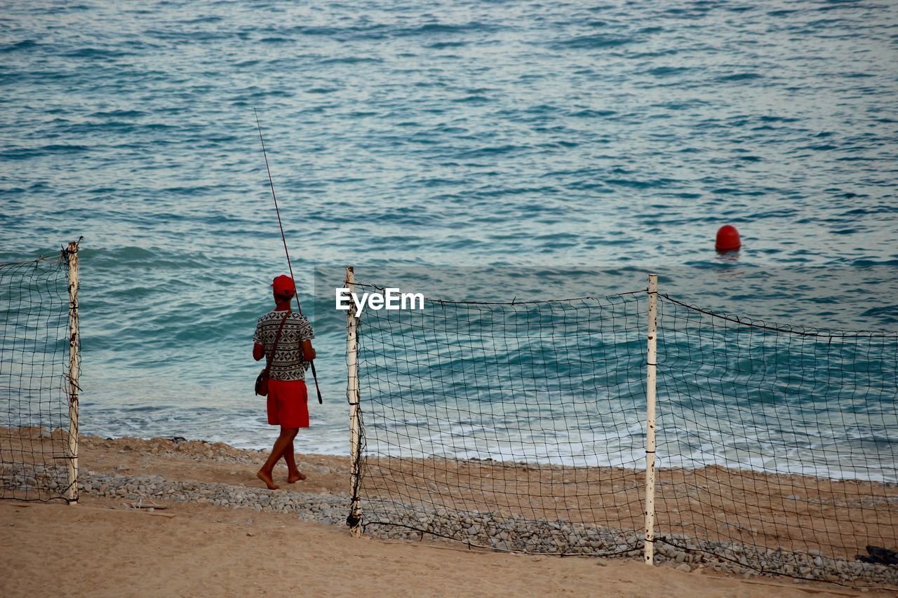 REAR VIEW OF MAN FISHING ON SEA SHORE