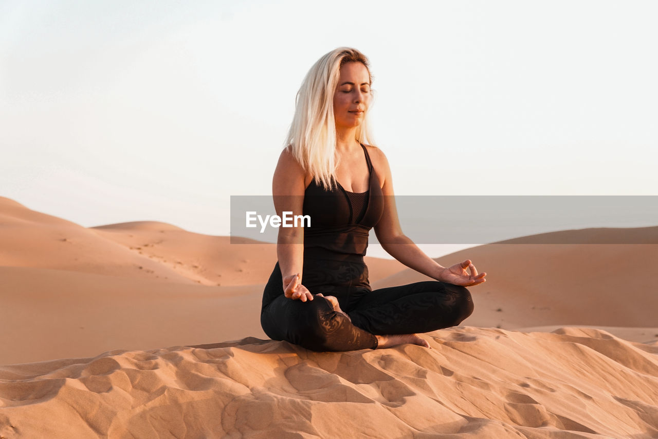 Young woman sitting on sand in desert against sky