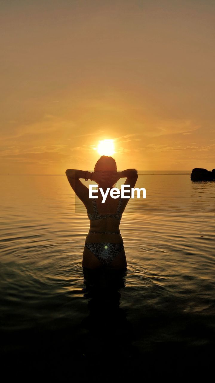 sunset, water, sky, one person, silhouette, nature, adult, reflection, sea, sunlight, women, beauty in nature, sun, leisure activity, vacation, tranquility, trip, holiday, lifestyles, orange color, standing, evening, back lit, land, relaxation, arm, beach, idyllic, dusk, outdoors, horizon over water, tranquil scene, scenics - nature, three quarter length, young adult, limb, horizon, summer, enjoyment, clothing, arms raised, happiness, rear view, female, zen-like