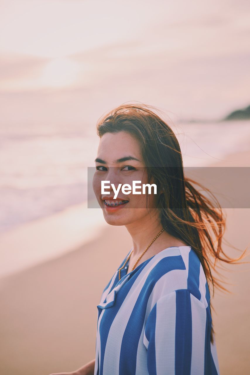 Side view portrait of smiling young woman standing at beach during sunset