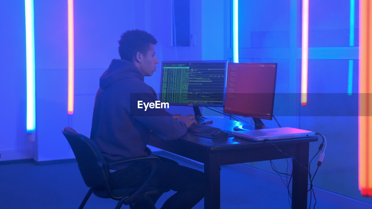 Young man in hood hacking by using computer while sitting at illuminated office
