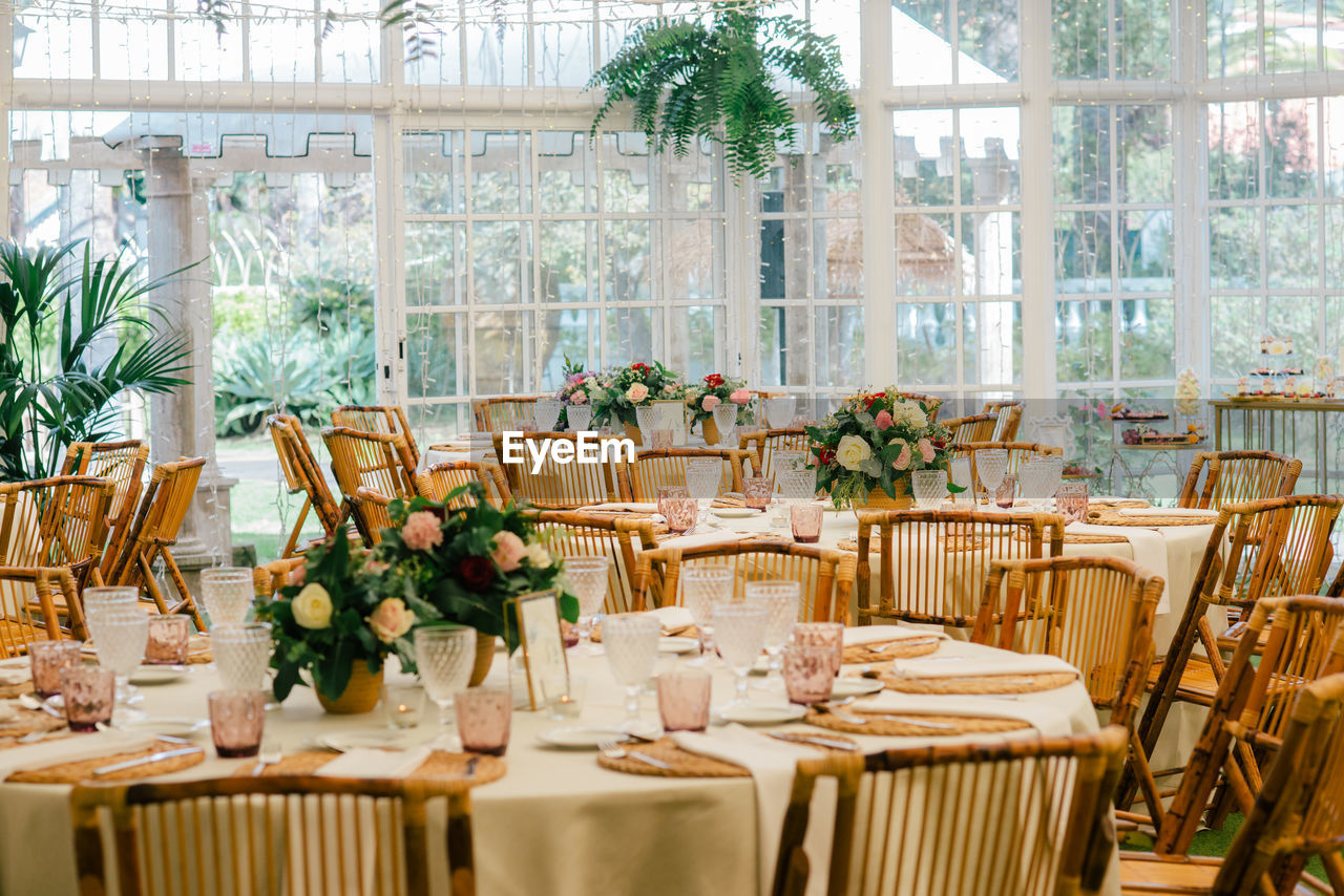 Big spacious room with festive decorated tables and wooden chairs under ceiling with green plants