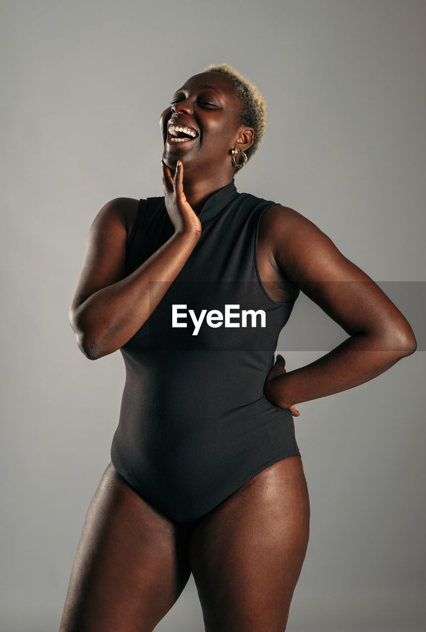 Cheerful african american curvy female in black bodysuit standing on gray background in studio and laughing
