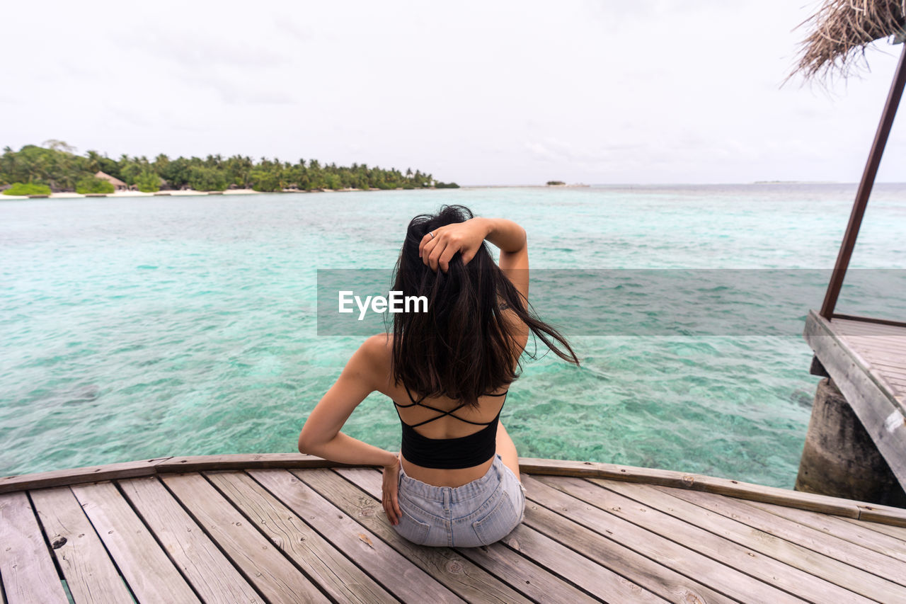 Back view of unrecognizable female in casual clothes sitting on wooden pier relaxing in maldives