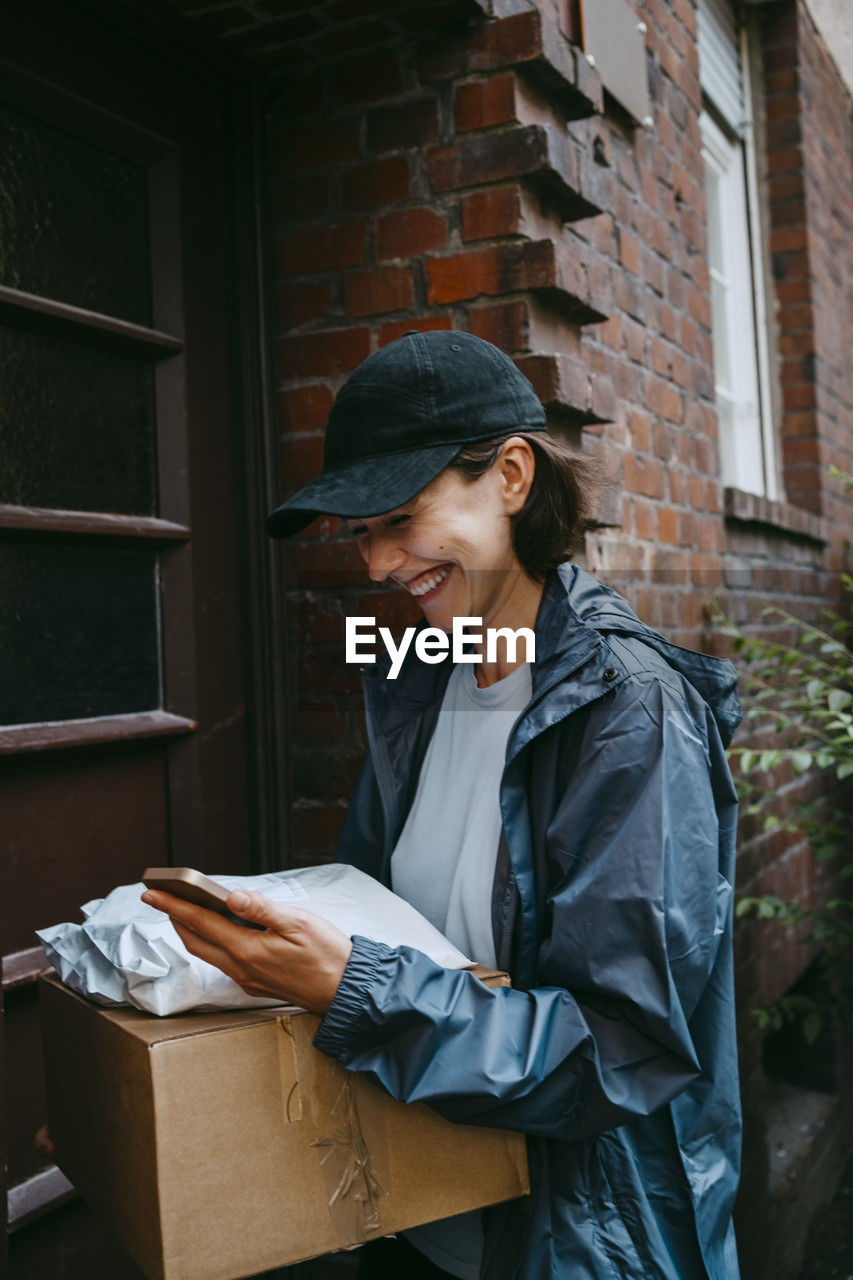 Happy female delivery person laughing while standing with parcel at doorstep