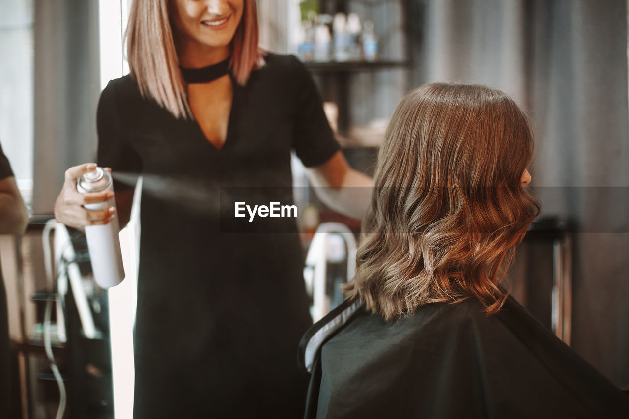 Smiling woman spraying beauty product on hair of customer at salon