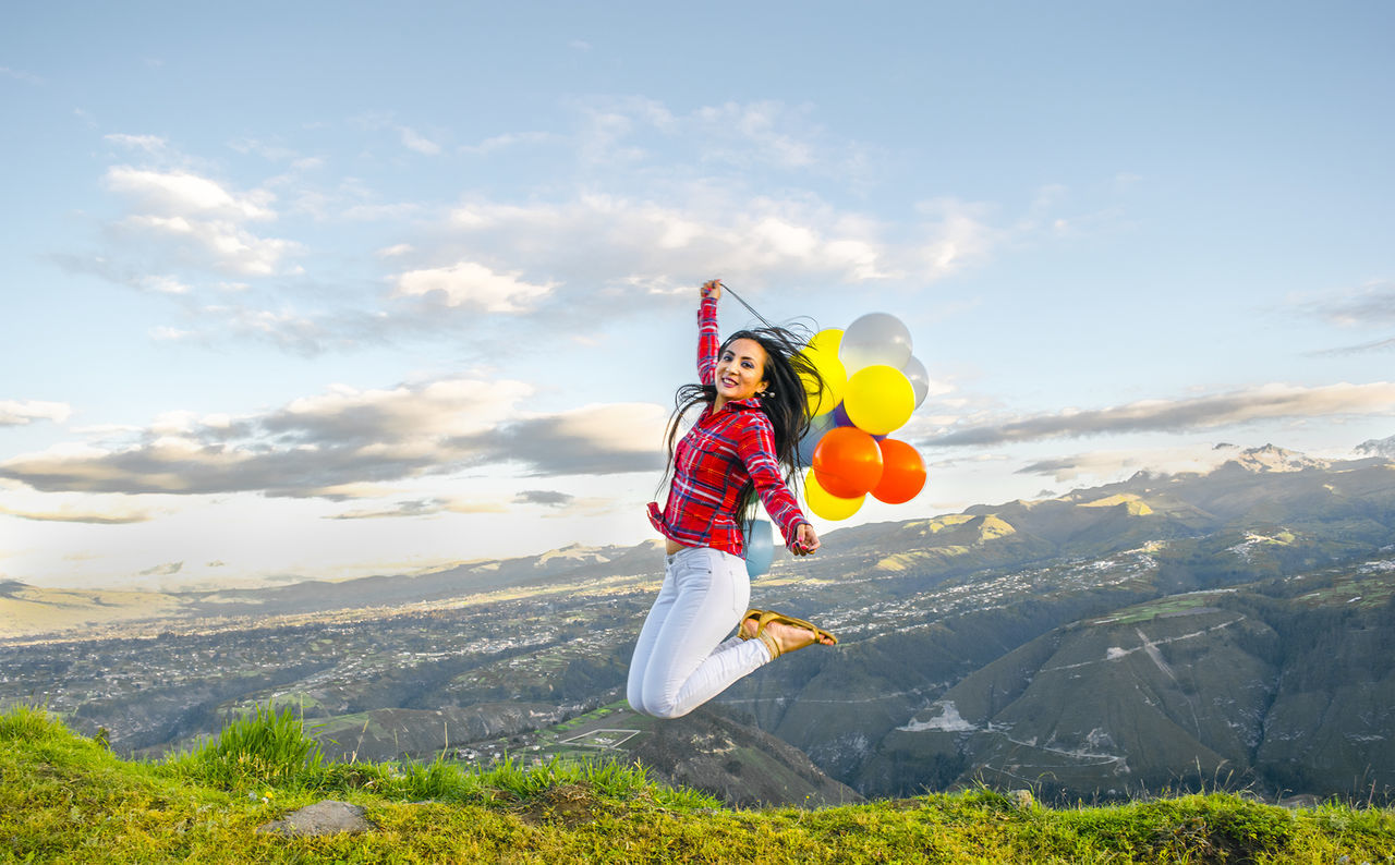 Portrait of excited woman holding balloons jumping at mountain peak against sky