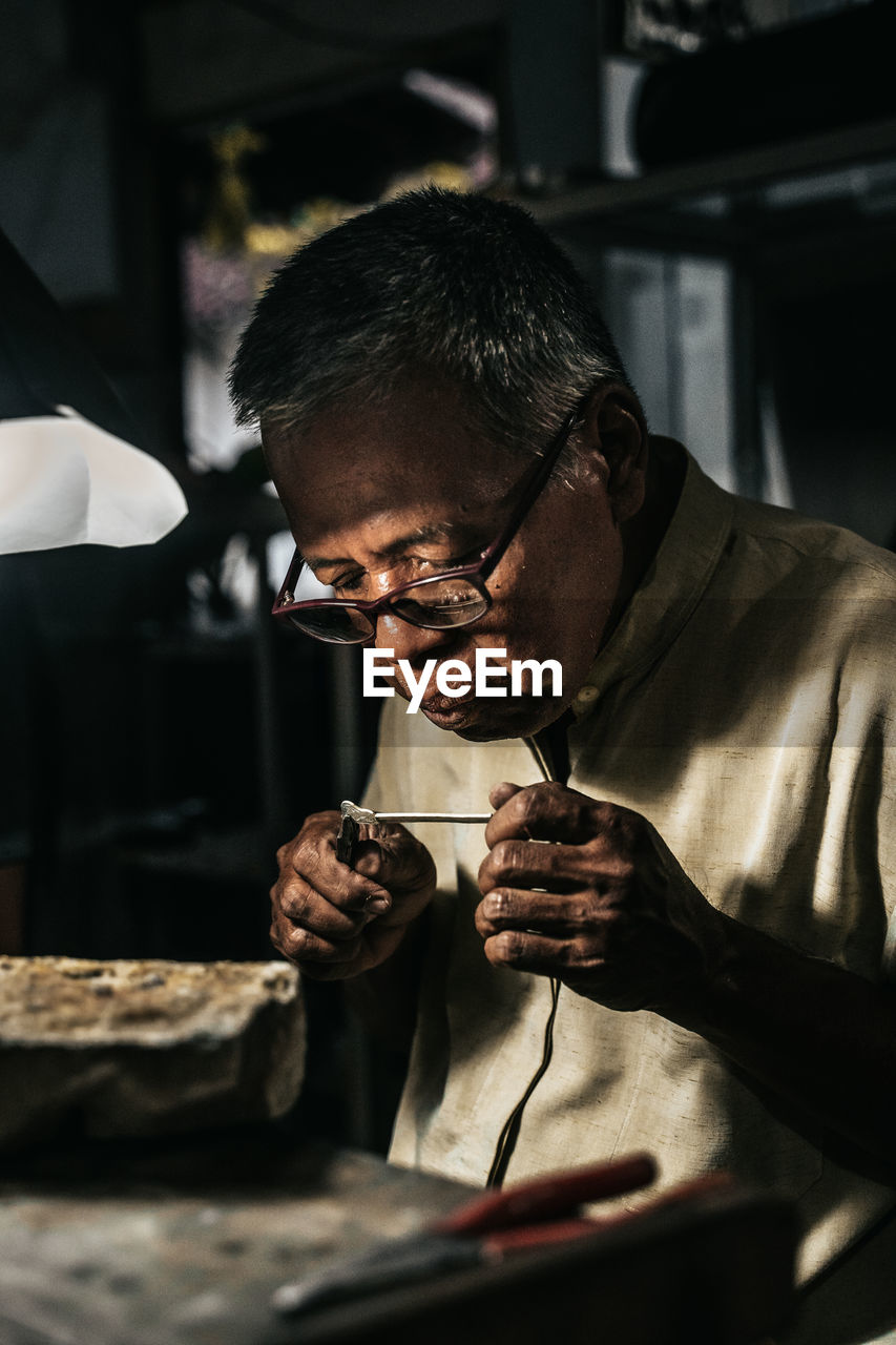 Concentrated senior african american male goldsmith in casual clothes and eyeglasses holding solder with pliers while creating jewelry in workshop