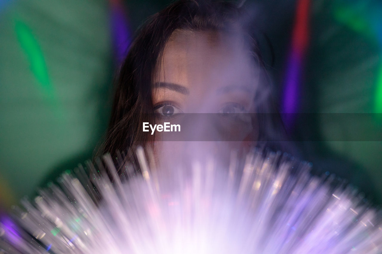 Close-up portrait of young woman holding illuminated fiber optic in darkroom