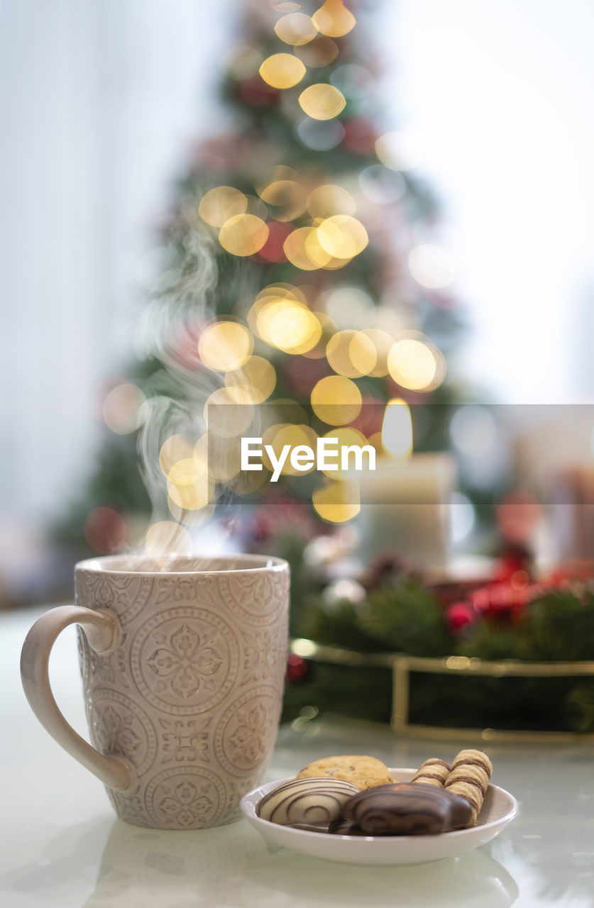 A cup of hot drink and in the background blurred candles and a christmas tree.